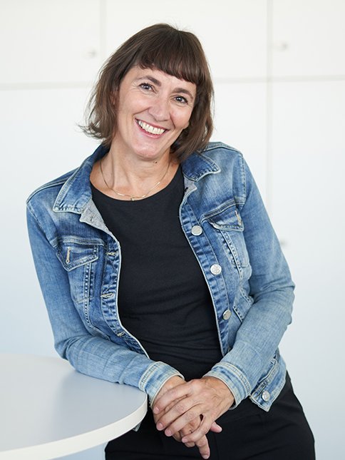 Eine Frau in einer Jeansjacke lehnt lächelnd an einem Stehtisch