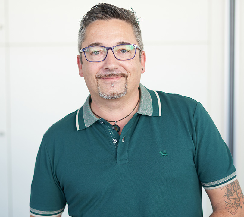Ein Mann mit kurzen Haaren, Brille und leichtem Bart trägt ein Polo-Shirt und hat ein Oberarmtatoo.